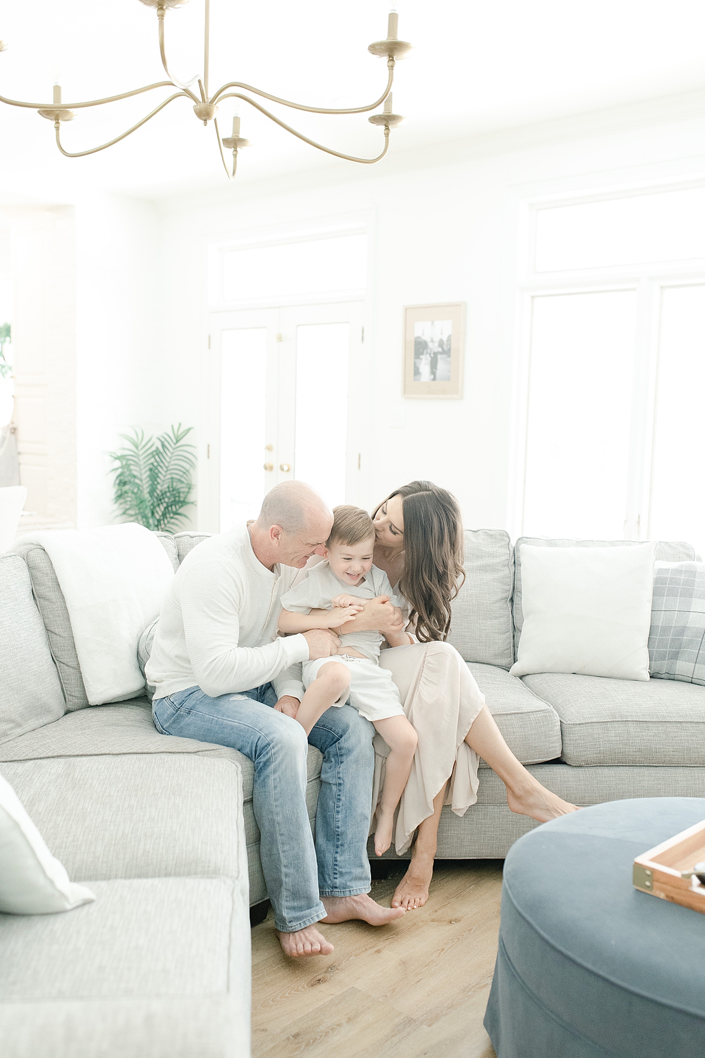 Mom and Dad kissing son while sitting on couch in living room. Photo by Little Sunshine Photography
