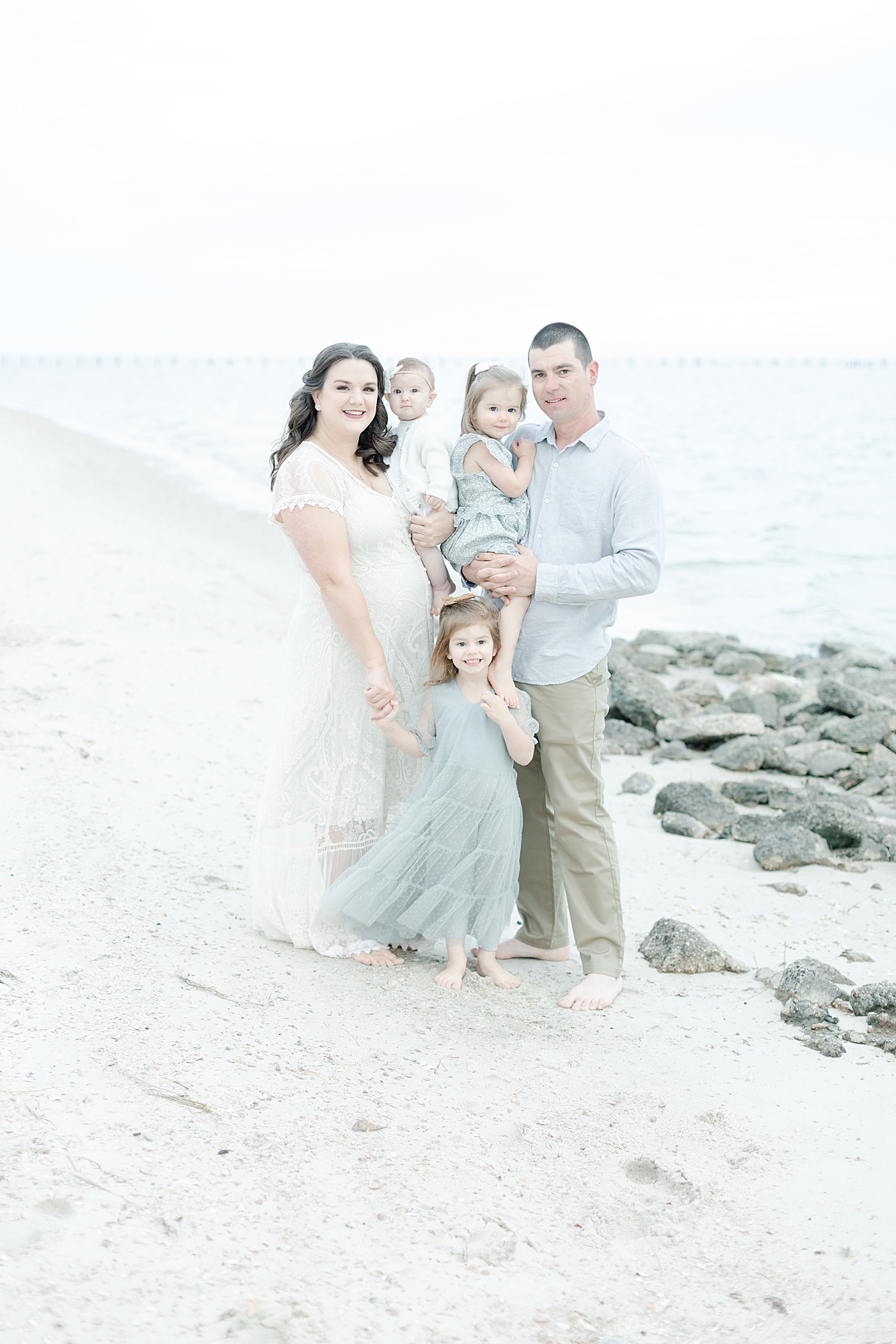 Family in white and blue standing near rocks on the beach | Photo by Little Sunshine Photography