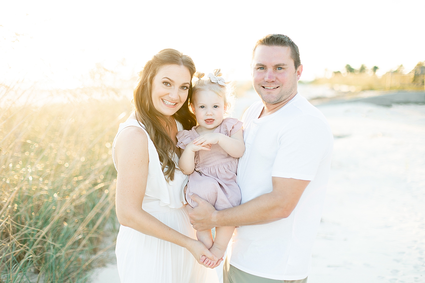Dad, mom, and toddler baby in pink smiling at the camera | Photo by Little Sunshine Photography