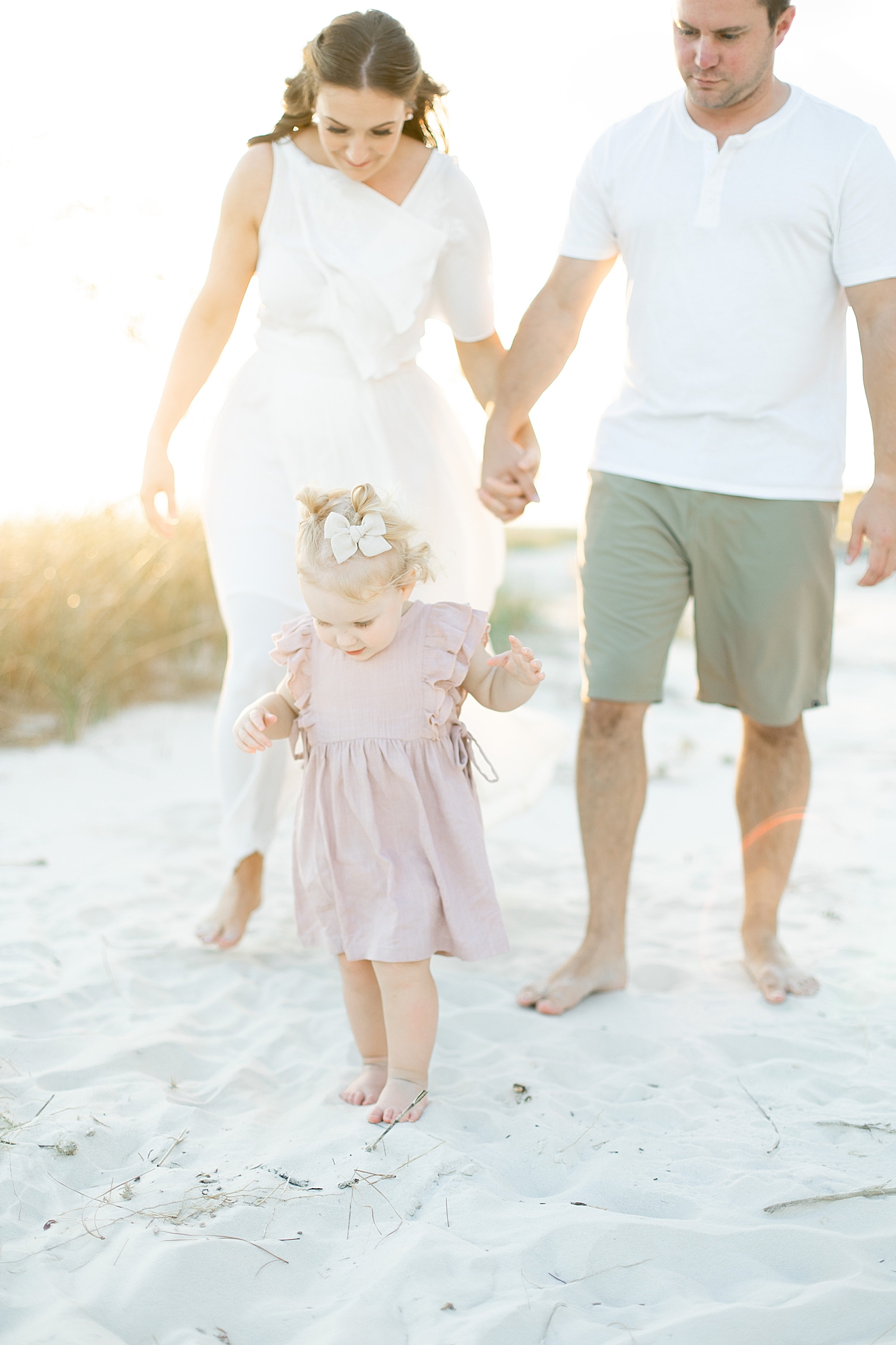 Mom and dad walking behind toddler baby girl on the beach | Photo by Little Sunshine Photography