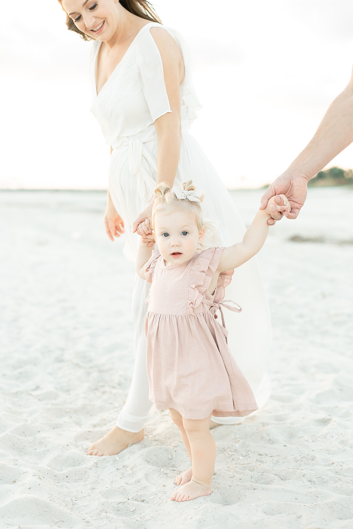 Toddler girl in pink dress walking with mom and dad on the beach | Photo by Little Sunshine Photography