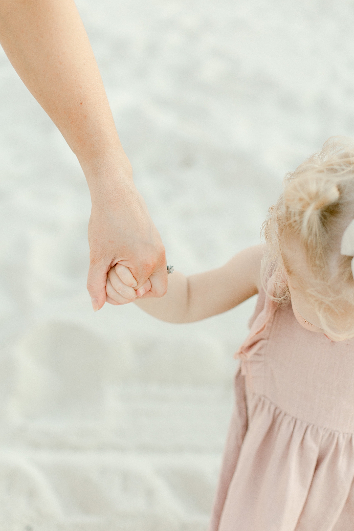 Toddler girl in pink bow holding moms hand on the beach | Photo by Little Sunshine Photography