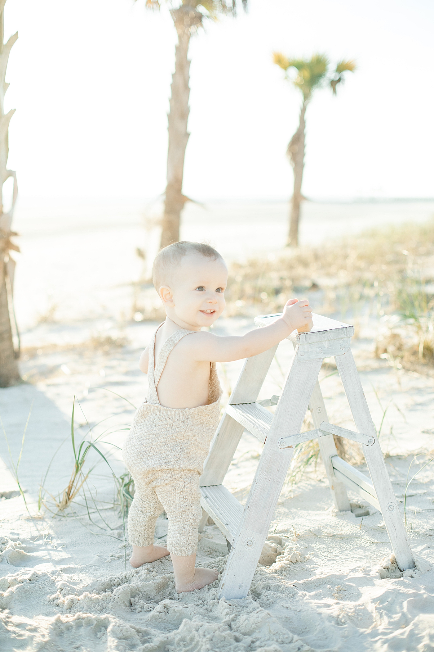  Baby boy standing on the beach with white ladder | Photo by Ocean Springs Family Photographer Little Sunshine Photography