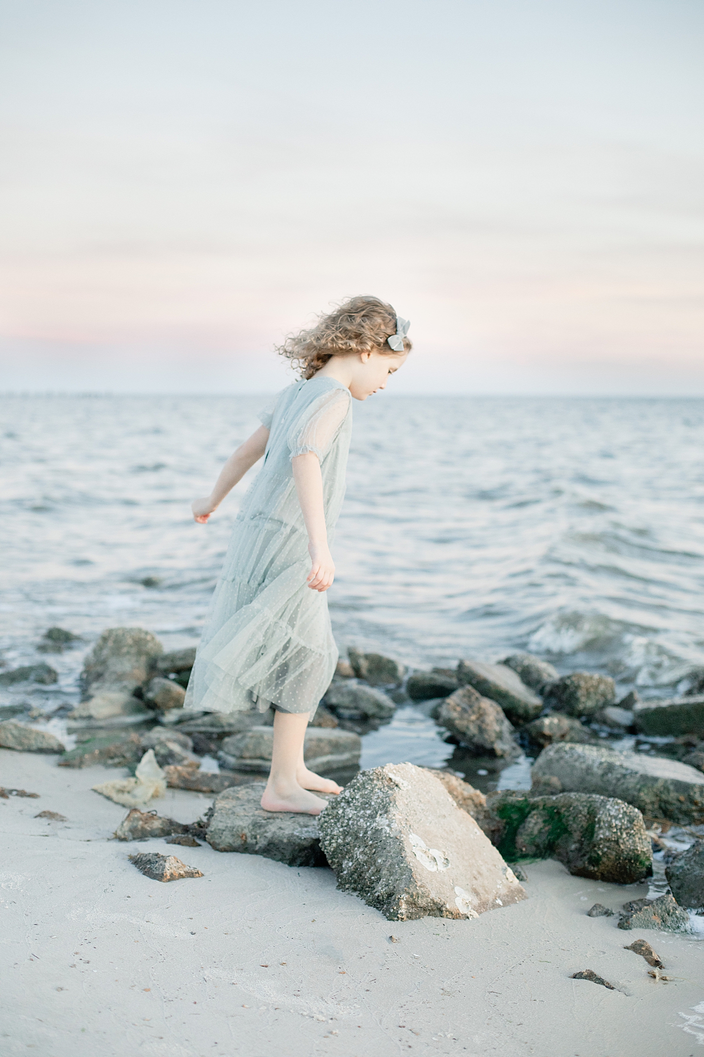 Little girl in gray dress walking on rocks at the beach | Photo by Little Sunshine Photography